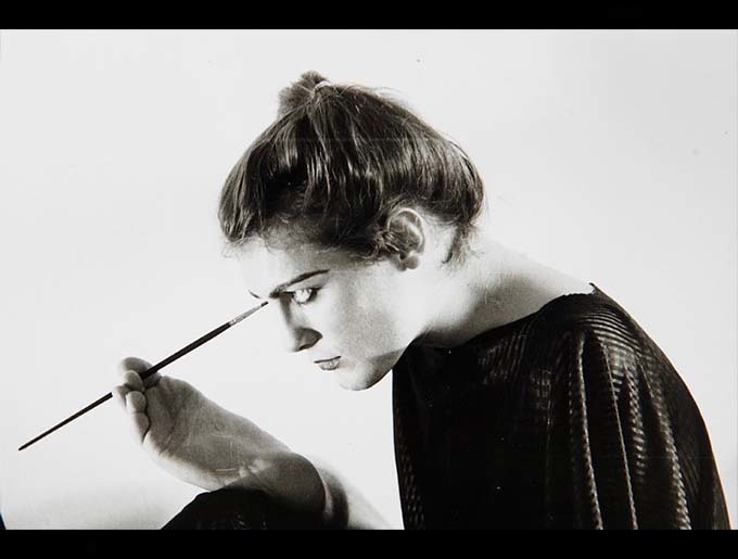 A black and white photograph of a light skinned woman applying her makeup using her foot. Her hair is in a bun and she is wearing a black flowing dress.