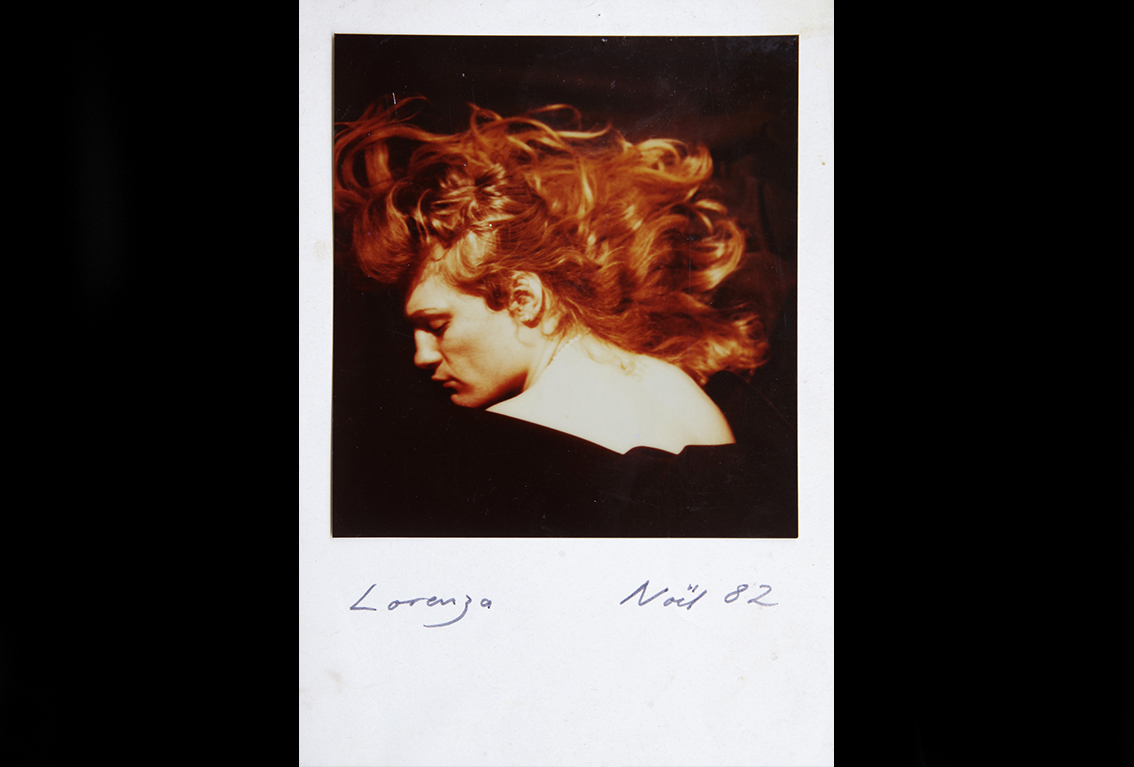 A polaroid photo of the back of a person looking over her shoulders. Red hair displayed and fanned out. The bottom left of the photo is signed Lorenza and the bottom right corner reads Noel 82.
