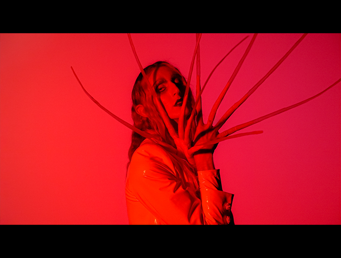 A figure with long hair is bathed in red light amidst a flat red background. They are shown from the waist up, gazing at the viewer, and with fingernails larger than their head covering their face.